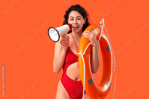 Beautiful young happy African-American female lifeguard with ring buoy and megaphone on orange background
