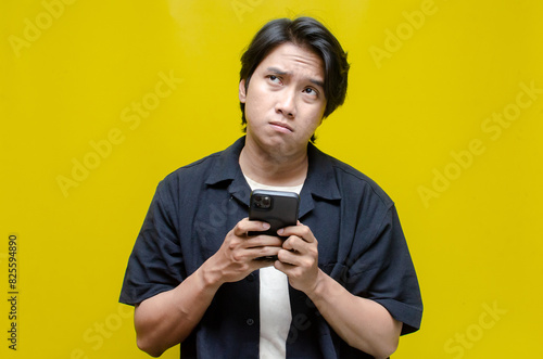 an overthinker young Asian man in pensive mood holding phone with sad and thoughtful expression. Asian man holding phone while questioning and wondering.