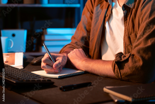 Cropped image of young businessman writing design thinking on paper for new project at neon light modern office. Thinking new next job sketching on notebook before sending present via email. Gusher.