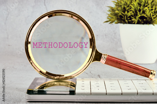 Business, study or activity concept. The word methodology written through a magnifying glass standing on a calculator on a gray background with a plant in the background without focus