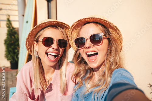 Two young beautiful smiling hipster female in trendy summer clothes. Sexy carefree women posing on street background. Positive models having fun, hugging at sunset. In hat and sunglasses. Take selfie