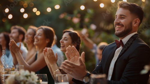 A smartly dressed man clapping with a smile at a well-lit outdoor evening celebration