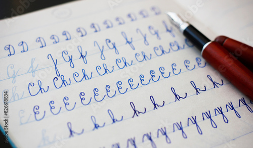 Close-up of uppercase and lowercase letters written in cursive with a fountain pen with blue ink in a lined notebook, a burgundy fountain pen in the background. Education and handwriting concept.