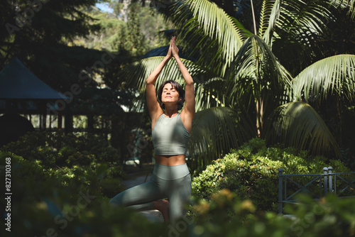 Fit sporty mindful young Hispanic woman meditating doing yoga breathing exercises with eyes closed feeling peace of mind, mental balance standing in green nature tropical park on sunny day.