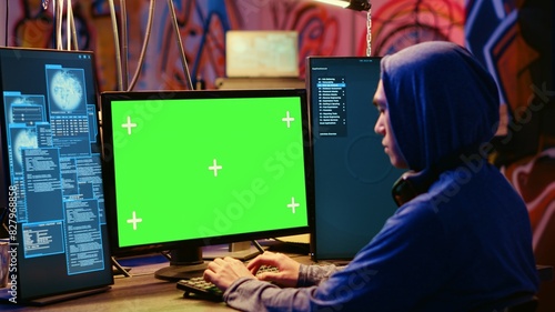 Asian hacker in hidden place with graffiti walls using green screen computer to deploy malware on unsecured devices, stealing sensitive data from unaware users online and selling it on black market
