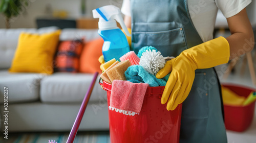 Woman wearing apron using yellow rubber gloves on her hands holding bucket containing cleaning supplies, plastic bottle spray, sponge, dust mop and colorful rags 