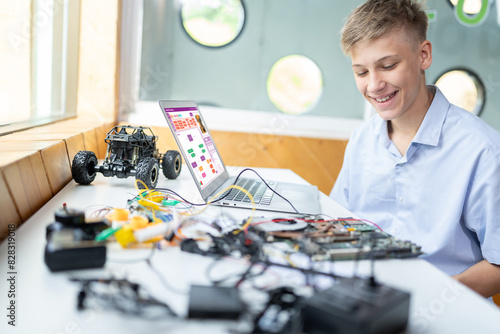 Blonde hair schoolboy in blue shirt watching motherboard while considering and smile in STEM class. On table put laptop, controller, electric wire, battery charger, and robotic vehicle. Edification.