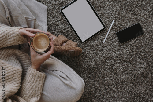 Cozy morning routine. Top view of woman wearing pastel creamy beige clothes sitting on fluffy grey carpet with cup of coffee. Tablet and mobile phone mock up with copy space