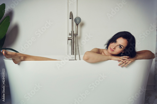 charming brunette woman in a black dress is sitting in the bathtub with her leg out