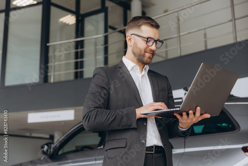 Modern bearded man in glasses and suit vehicle sales consultant using laptop inside car dealership