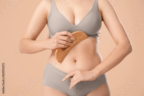 Young woman in underwear massaging her belly with wooden scraper on beige background, closeup