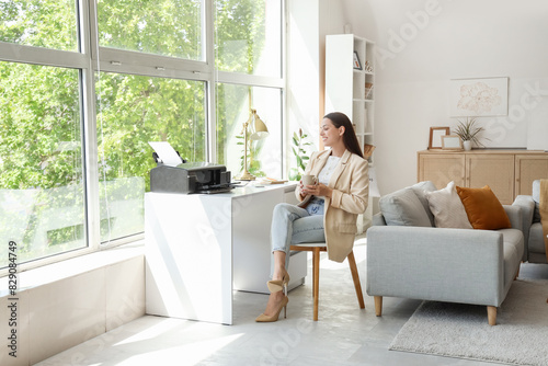 Young businesswoman with cup of coffee and printer on table in office