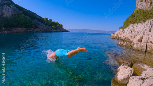 Young guy in shorts splashes into crystal blue sea from raised rock on the shore. Refreshing dive of a young man into ocean from a rocky coast, embodying freedom and joy of carefree summer adventures