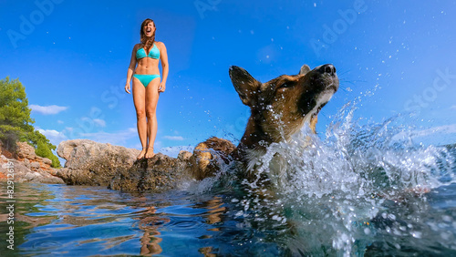 LOW ANGLE VIEW: Lady in bikini stands on sea rock when her dog jumps into water. Upon landing in crystal blue sea, adorable brown doggo makes a big splash. Active summer holidays at Dalmatian seaside.