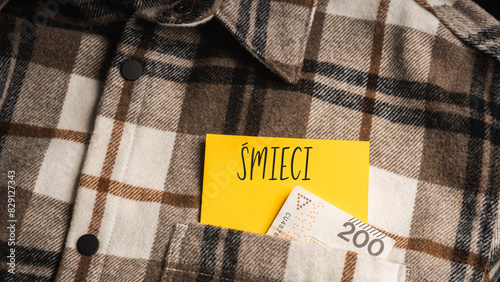 Yellow card with a handwritten inscription "Śmieci", protruding from a brown plaid shirt, next to Polish banknotes PLN (selective focus), translation: Garbage 