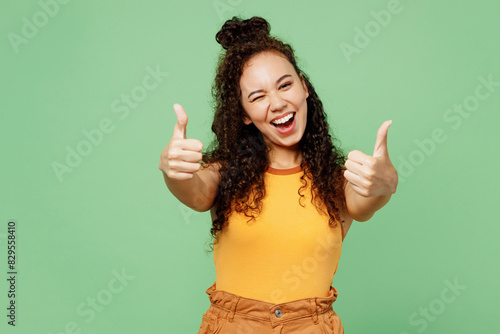 Young happy woman of African American ethnicity wear yellow tank shirt top showing thumb up like gesture blink eye isolated on plain pastel light green background studio portrait. Lifestyle concept.