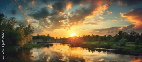 A picturesque sunset with the sun setting over the horizon casting golden rays on a bridge over a river highlighting a lush green clearing with tall trees under a sky painted with colorful clouds Copy