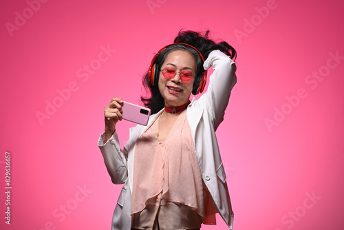 Cheerful elderly woman with headphones enjoying music dancing over pink background