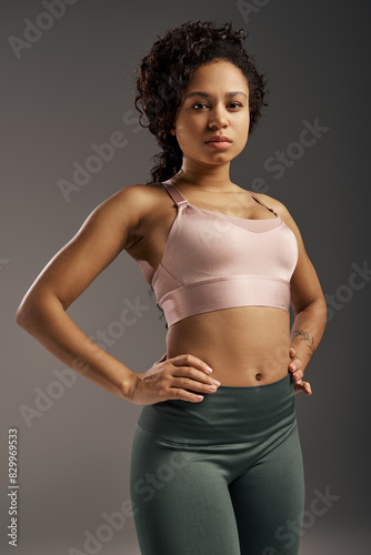 Curly African American sportswoman in pink sports bra and green leggings working out in studio with grey background.