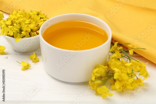 Rapeseed oil in bowl and beautiful yellow flowers on white wooden table