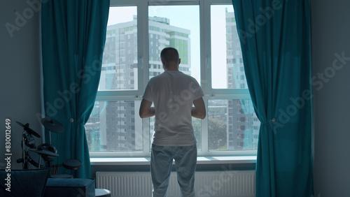 Rear view of a pensive man standing at home by the window and looking at the city. Media. Man in home clothes spending time in reflection in his bedroom.