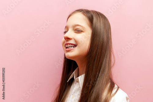 cheerful teenage girl with braces winks and hints on pink isolated background, happy child blinks eye and jokes