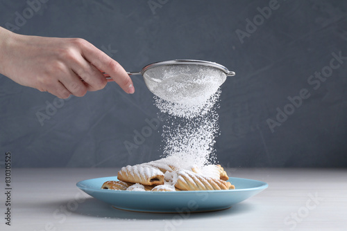 Woman with sieve sprinkling powdered sugar onto cookies at white wooden table, closeup