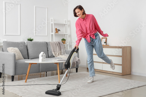 Happy young woman cleaning carpet with handheld vacuum cleaner in living room