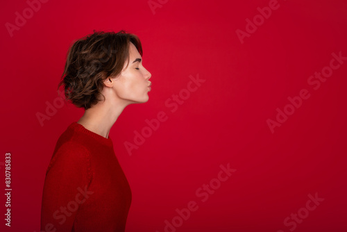 Side view profile image of beautiful young woman with short curly hairstyle in red bright sweater, she imagines kissing someone isolated on the red vivid wall