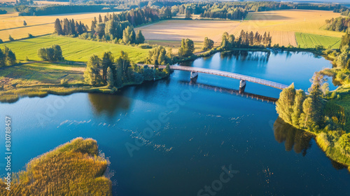 Aerial view of a bridge spanning a blue lake on a sunny summer day in rural Finland, surrounded by lush green and golden yellow fields, creating a picturesque countryside scene.