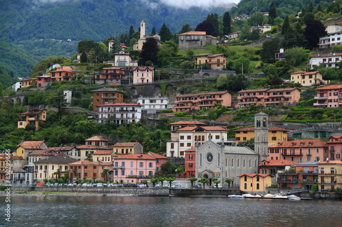 A landscape with a view of Lake Como and the Chiesa Santissima Trinita church in the town of Argegno with the mountains in the background covered with greenery and partly with clouds