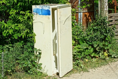 one old dirty closed discarded rusty white electric closed iron refrigerator stands on the ground near green bushes during the day on the street