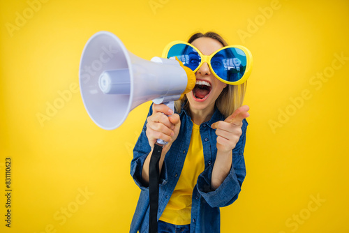 happy vivid young woman in funny large sunglasses scream in megaphone announces discounts sale. Hurry up isolated on yellow background studio portrait
