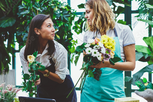 Female colleagues working as florist in botanic orangery cooperating during creating floral composition,young woman in apron assist professional owner of flower store making bouquet for clients order