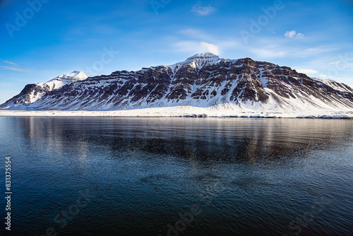 2023-12-31 A RAGGED MOUNTAIN PARTIALLY COVERED WITH SNOW WITH A CLAM ARCTIC OCEANAND A NICE BLUE SKY NEAR SVALBARD NORWAY IN THE ARCTIC