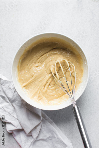 Overhead view of vanilla cake batter being whisked in a white ceramic bowl, flatlay of cake batter being mixed, process of making cake