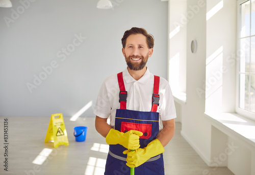 Janitor from cleaning service at work. Cleaning company worker helps keep indoor space clean and sanitary. Happy man in workwear standing inside building, holding mop, looking at camera and smiling