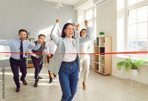 Happy excited woman employee running to red ribbon celebrating crossing finishing line in office with people colleagues in background. Business competition, success and teamwork concept.