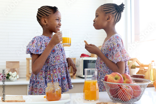African twin girl sister with curly hair braid African hairstyle eat bread toast with jam and drink orange juice at kitchen. Happy smiling kid sibling have breakfast together. Cute children in family.