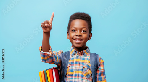 Smiling boy in a plaid shirt with a backpack and books, pointing upwards against a blue background.