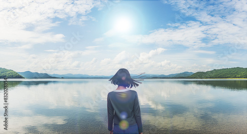 風に髪をなびかせ自然あふれる湖畔の岸に佇む女性の後ろ姿 夏の太陽の強い日差しとレンズフレア 青空と雲を反射する湖面 遠くの山々に海岸 アウトドア・バカンス・夏休み・キャンプ・旅行・観光・リラクゼーションのイメージ