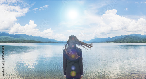 風に髪をなびかせ自然あふれる湖畔の岸に佇むスーツ姿の女性の後ろ姿 夏の太陽の強い日差しとレンズフレア 青空と雲を反射する湖面 遠くの山々に海岸 アウトドア・バカンス・夏休み・キャンプ・旅行・観光・リラクゼーションのイメージ