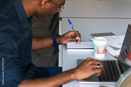 Man working from office taking reading and writing notes in note pad while working on laptop computer casual businessman entrepreneur focused doing research