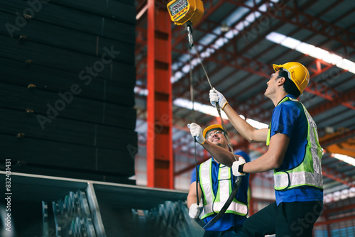 Young caucasian industrial worker holds belt for lift heavy load on hoist of crane in steel factory.