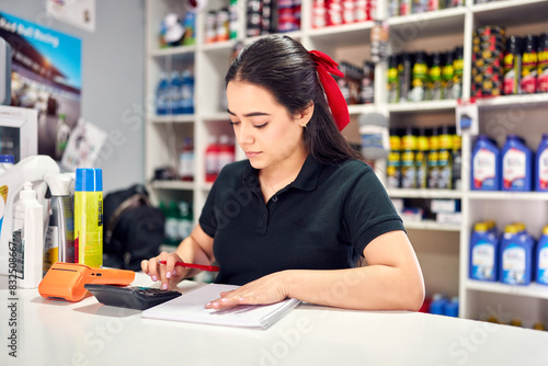 Portrait young latin woman behind counter working, using calculator and writing in small auto parts store