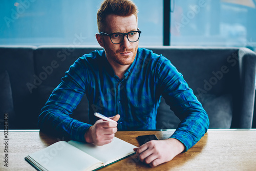 Handsome concentrated skiled student in optical eyeglasses thinking on writing interesting essay in notebook.Smart young man in spectacles pondering on creating new business plan for star tup project