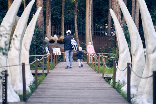 A father with two children enters Jurassic Park over a wooden bridge. At the entrance of the park there are white dinosaur tusks, with space for copying. High quality photo