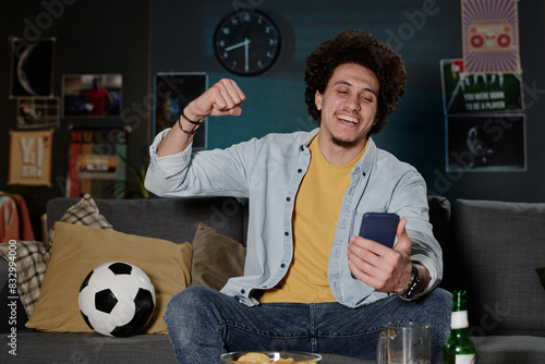 Medium shot of joyful biracial guy sitting on sofa late in evening watching soccer match online on smartphone, copy space