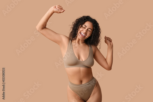 Beautiful young happy African-American woman in underwear dancing on beige background