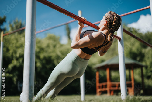 Woman practicing calisthenics at an urban park, doing pull-ups on a sunny day. Demonstrating strength, fitness, and an active lifestyle in outdoor environment.